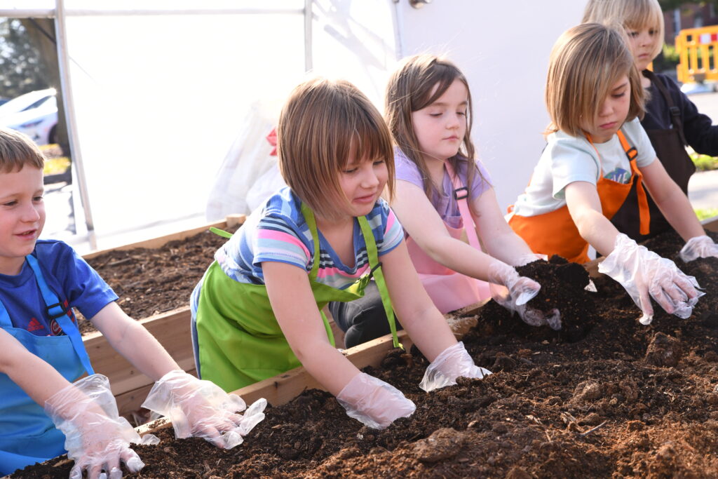 Five children wearing plastic gloves touch dirt.