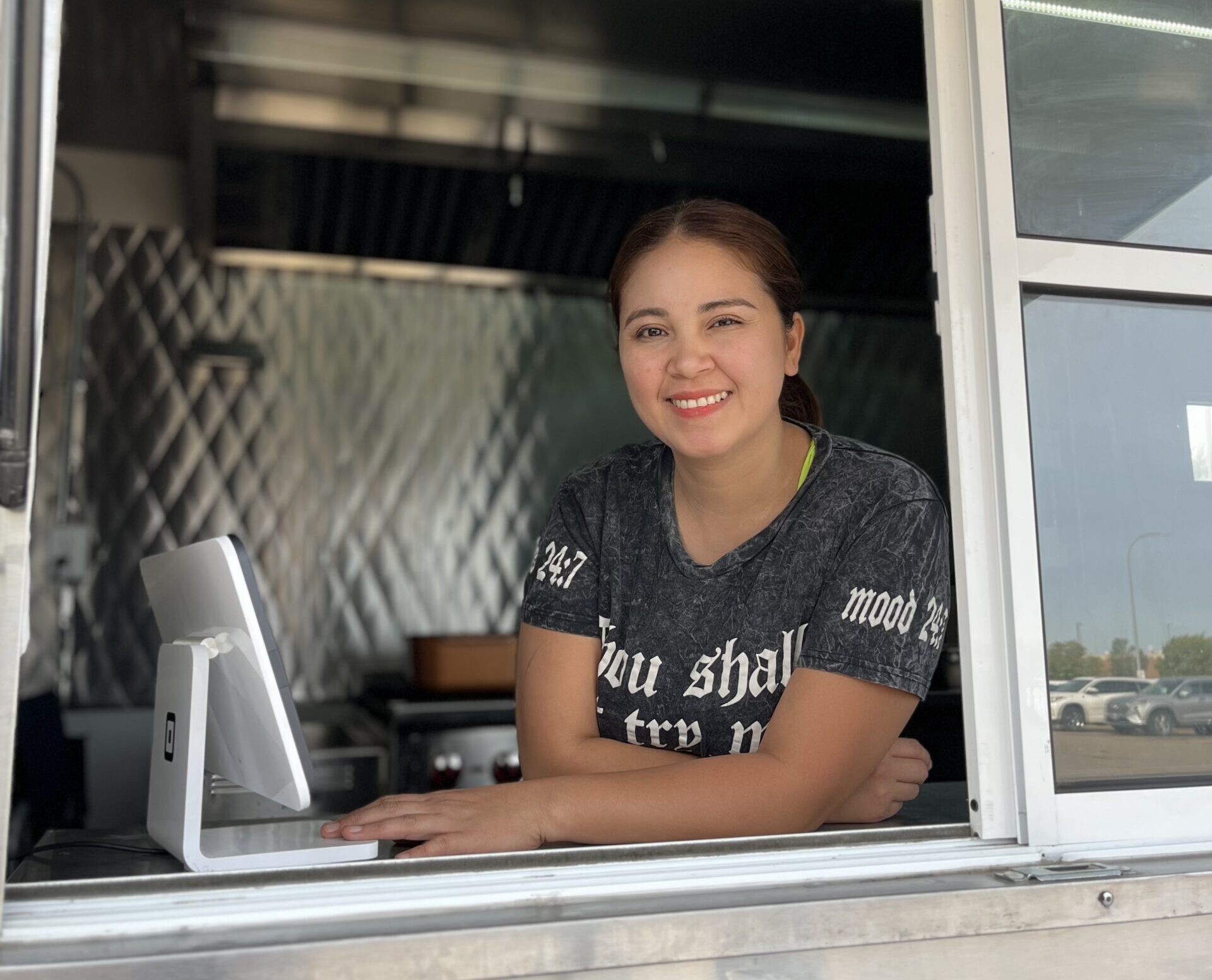 A person of medium skin tone looking out the window of a food truck and smiling. They have their dark hair tied back in pony and are wearing a dark grey tshirt with lettering on it.