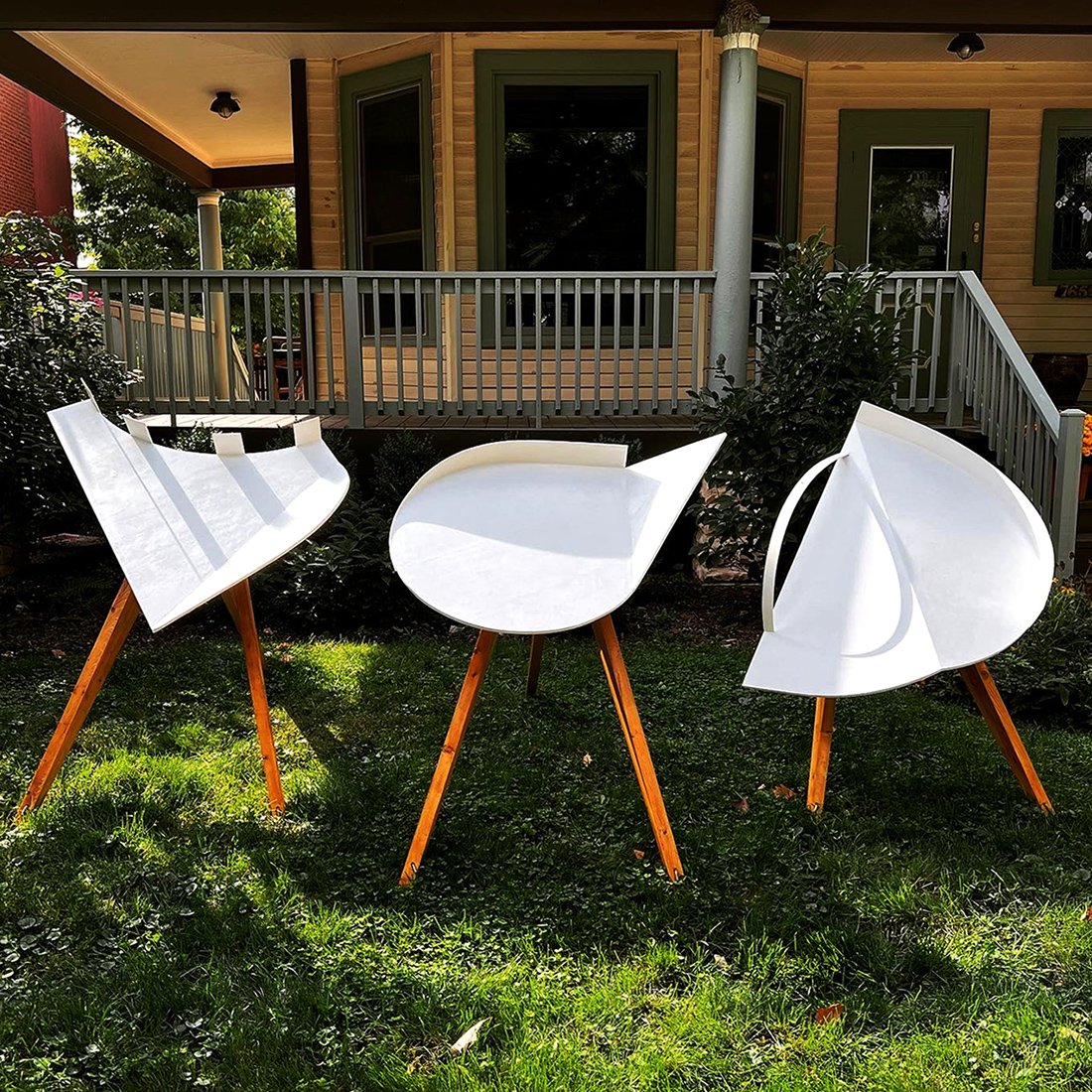 Three geometric sculptures in white with wooden legs placed outside in a yard of a house.