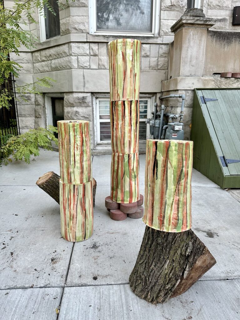 An artwork made of cut tree stumps, circular stone pavers, and cylindrical objects. They are stacked on top of each other and are on the sidewalk.