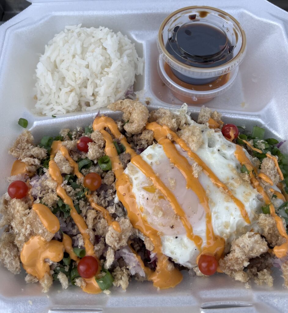 A takeout container with a small heap of rice and a large portion of a dish topped with a fried egg, crispy pork skin, cherry tomatoes, and a yellow sauce.