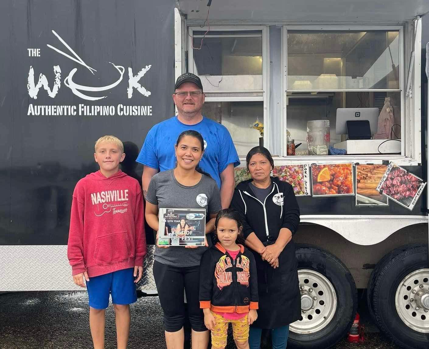 Three adults and two children posing in front of a black food truck. The truck has a sign that reads "the wok, authentic filipino cuisine"