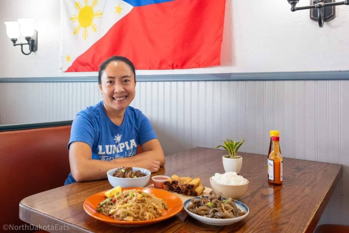 A person of medium skin tone sitting at a table and smiling. There are plates and bowls of food in front of her on the table.