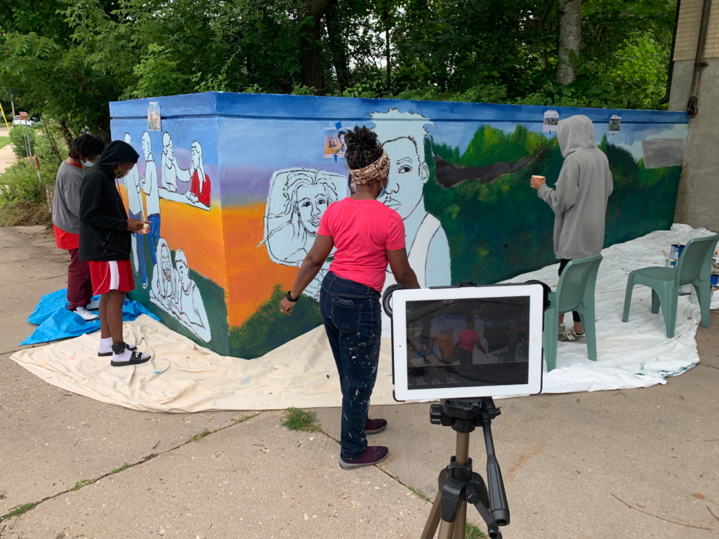 Four people stand by an exterior wall as they paint a large mural.