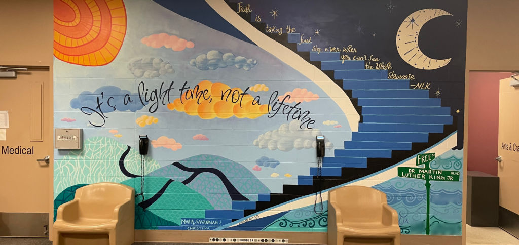 A wall with a large mural painting depicting stairs, the sun, the moon, and a skyscape.