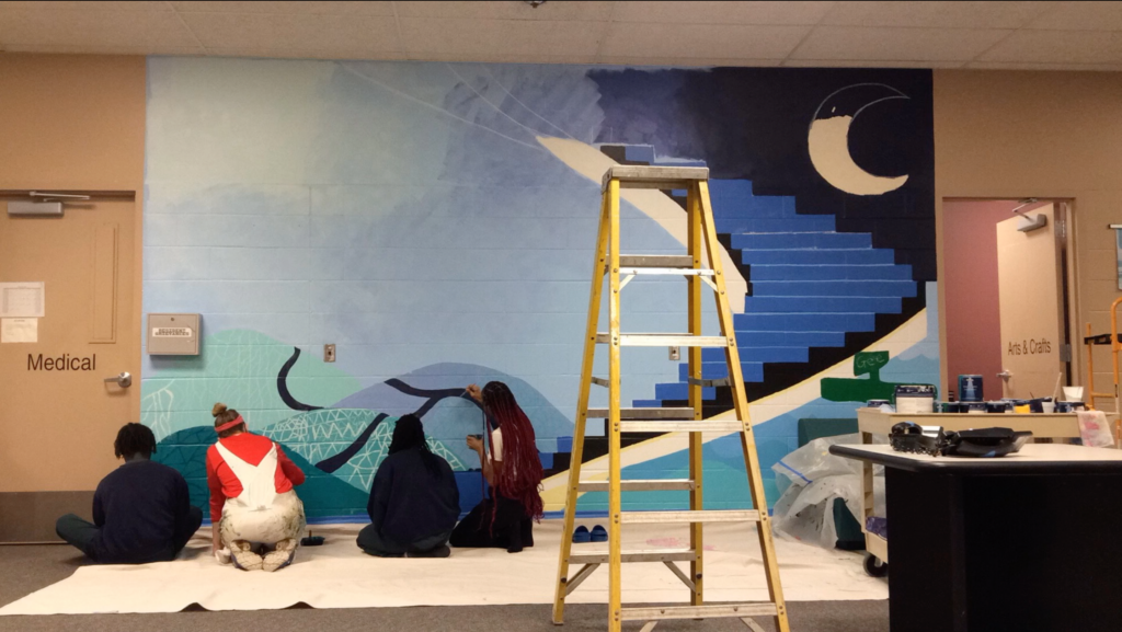 Four young people sitting or kneeling on the floor as they paint a large wall mural.