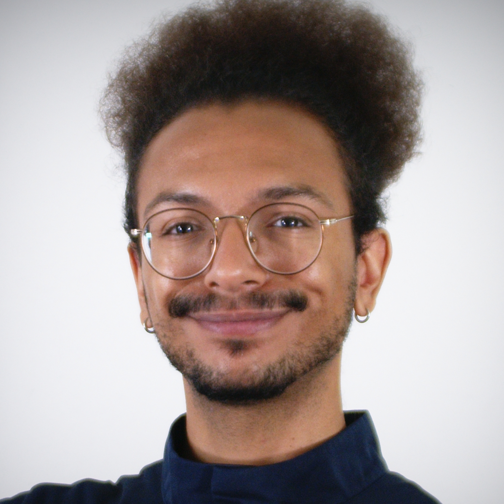 A person of medium skin tone, dark curly hair, and a mustache, wearing wire-rimmed glasses and a black turtleneck.