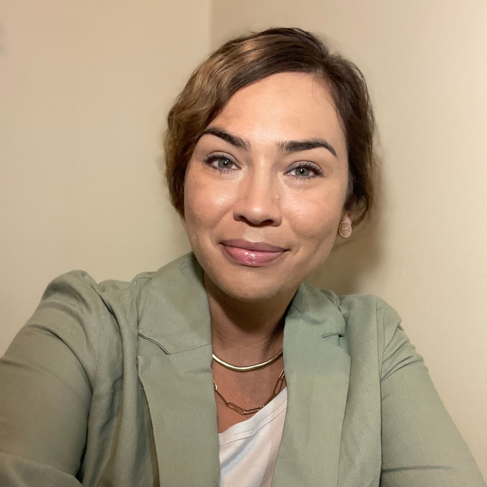 A person of medium light skin tone with brown hair in a low ponytail, wearing a white blouse and green blazer.