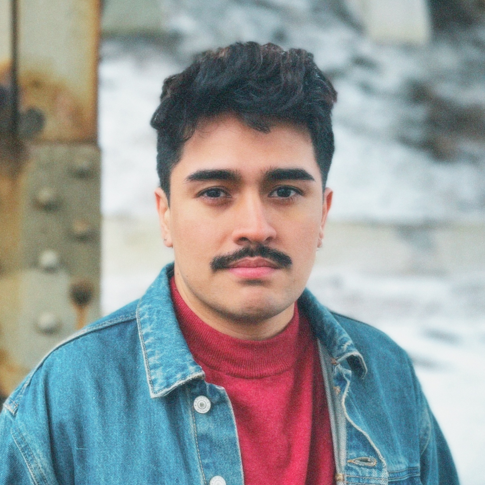 A person of medium-light skin tone with short black hair and a mustache wearing a red shirt and denim jacket.