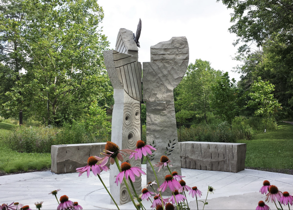 Two tall stone sculptures next to each other outdoors. They are surrounded by greenery.