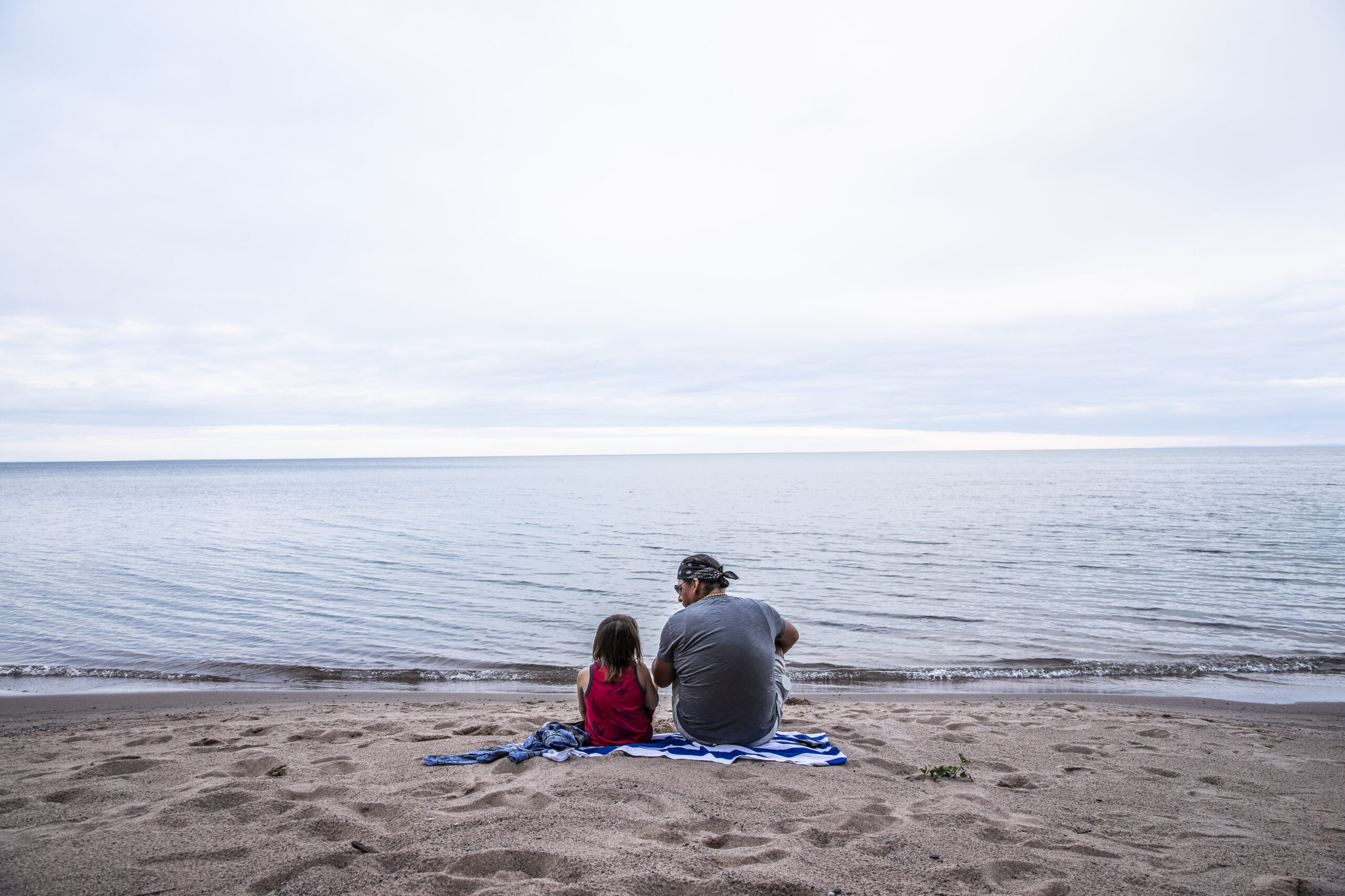 A man and a young boy sit on a beach overlooking water.