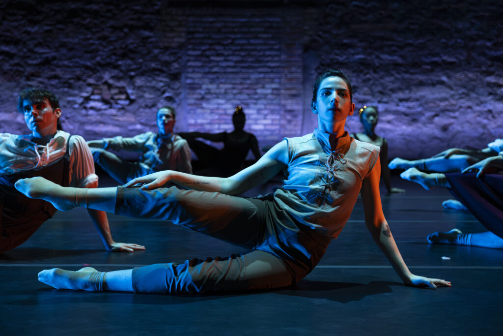 A group of dancers bathed in blue light extending one leg out horizontal to the ground while sitting