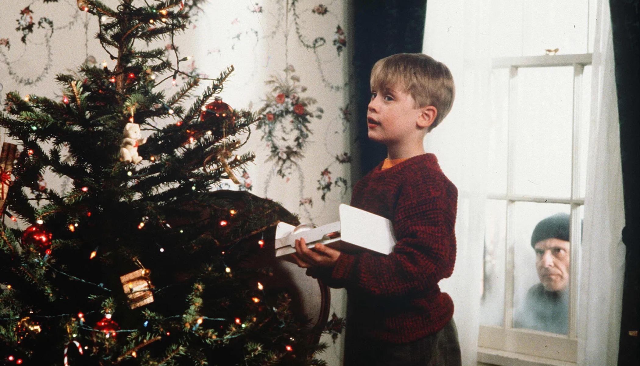 A screengrab from the film Home Alone of a child standing in front of a Christmas tree, while a man looks in the window behind him.