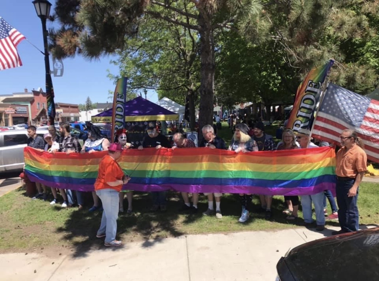 A group of people hold up a long rainbow colored flag. They are outdoors with trees, roads, and cars around them.