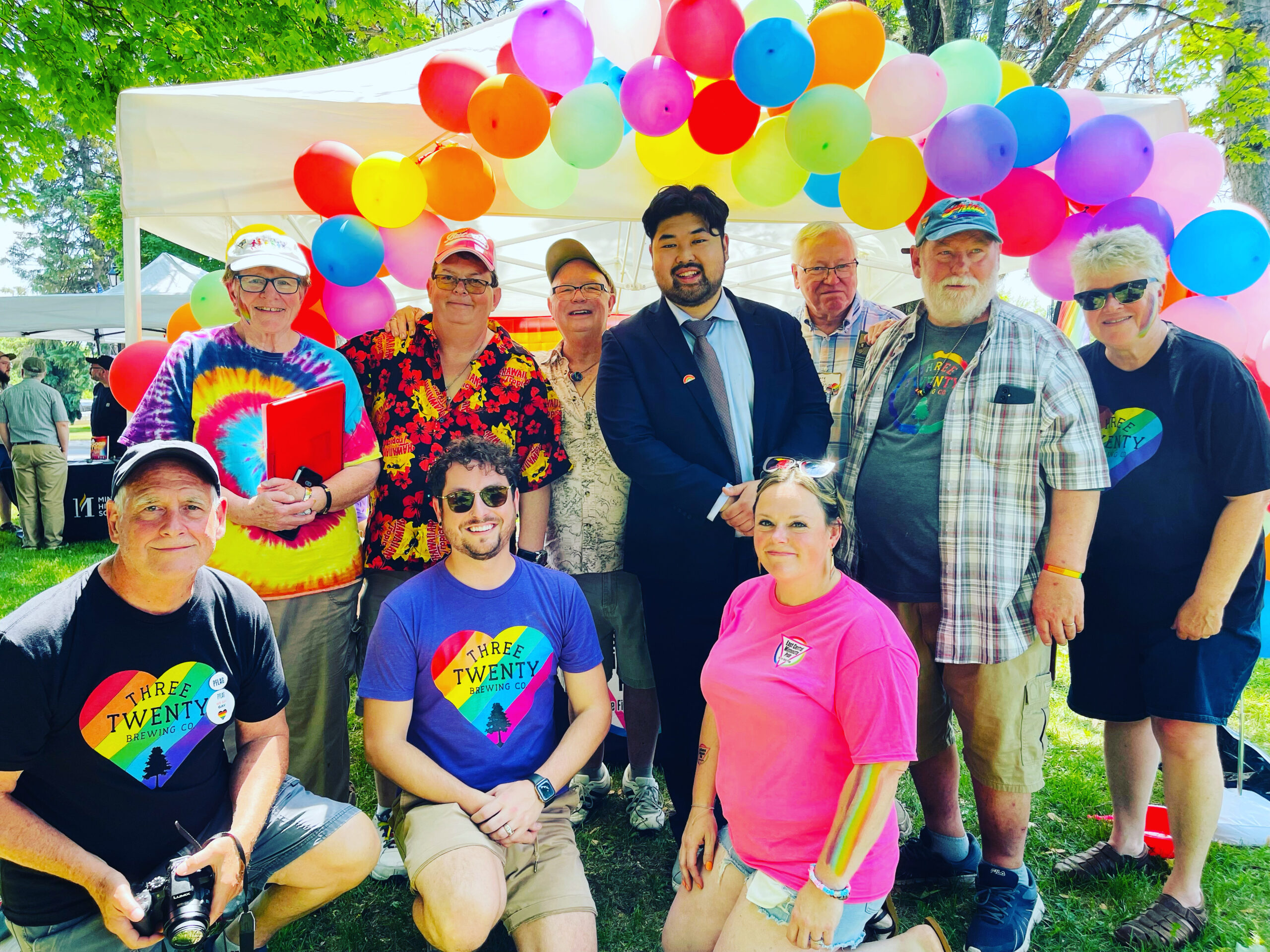 A group of people posing under a colorful balloon archway outdoors. Some people are kneeling, while others are standing.