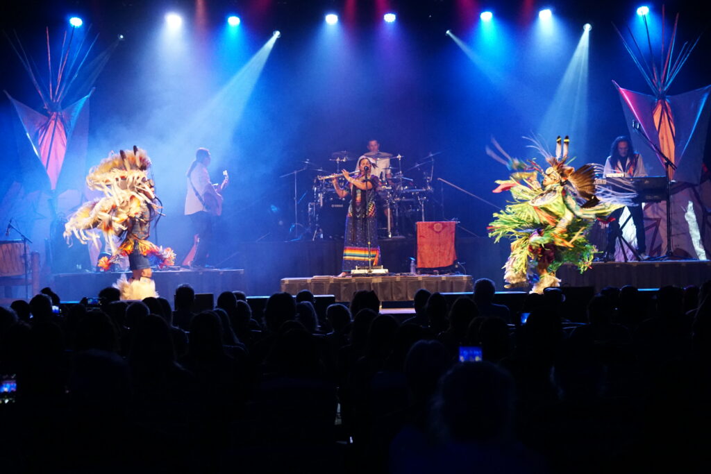 A guitarist, drummer, keyboard, and flutist performing on a stage with two dancers wearing traditional Native regalia.