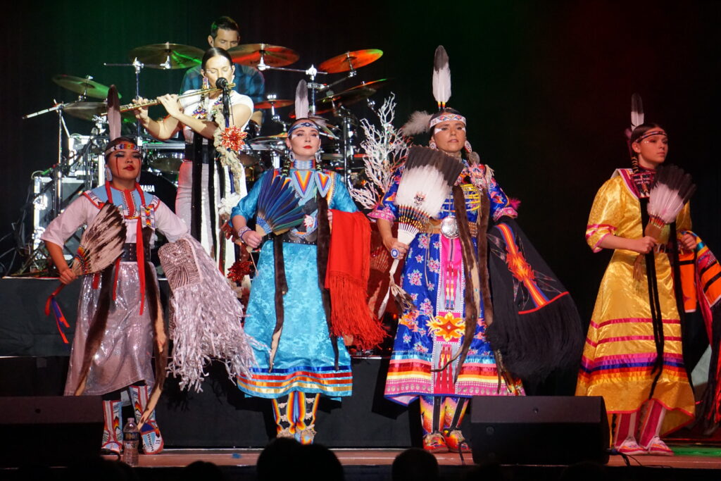 Four dancers in traditional Native regalia standing on stage in front of a flutist and drummer.