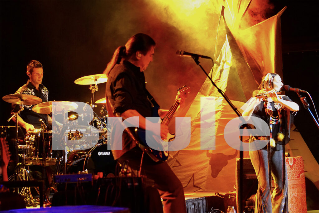 A guitarist, drummer, and flutist performing on stage in front of a large tipi, with overlaid text reading, "Brulé."