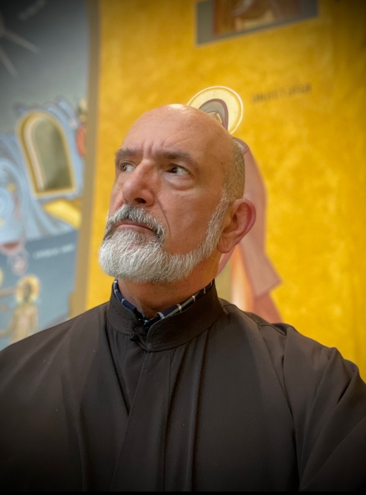 A bald man with a light skin tone and a white beard and mustache looks into the top left corner of the frame, his brow slightly furrowed and his gaze intense. He wears a black robe with a short collar. Behind him is religious art painted in bright gold, yellow, pink, and blue. The figures are mostly obscured by the man's body.