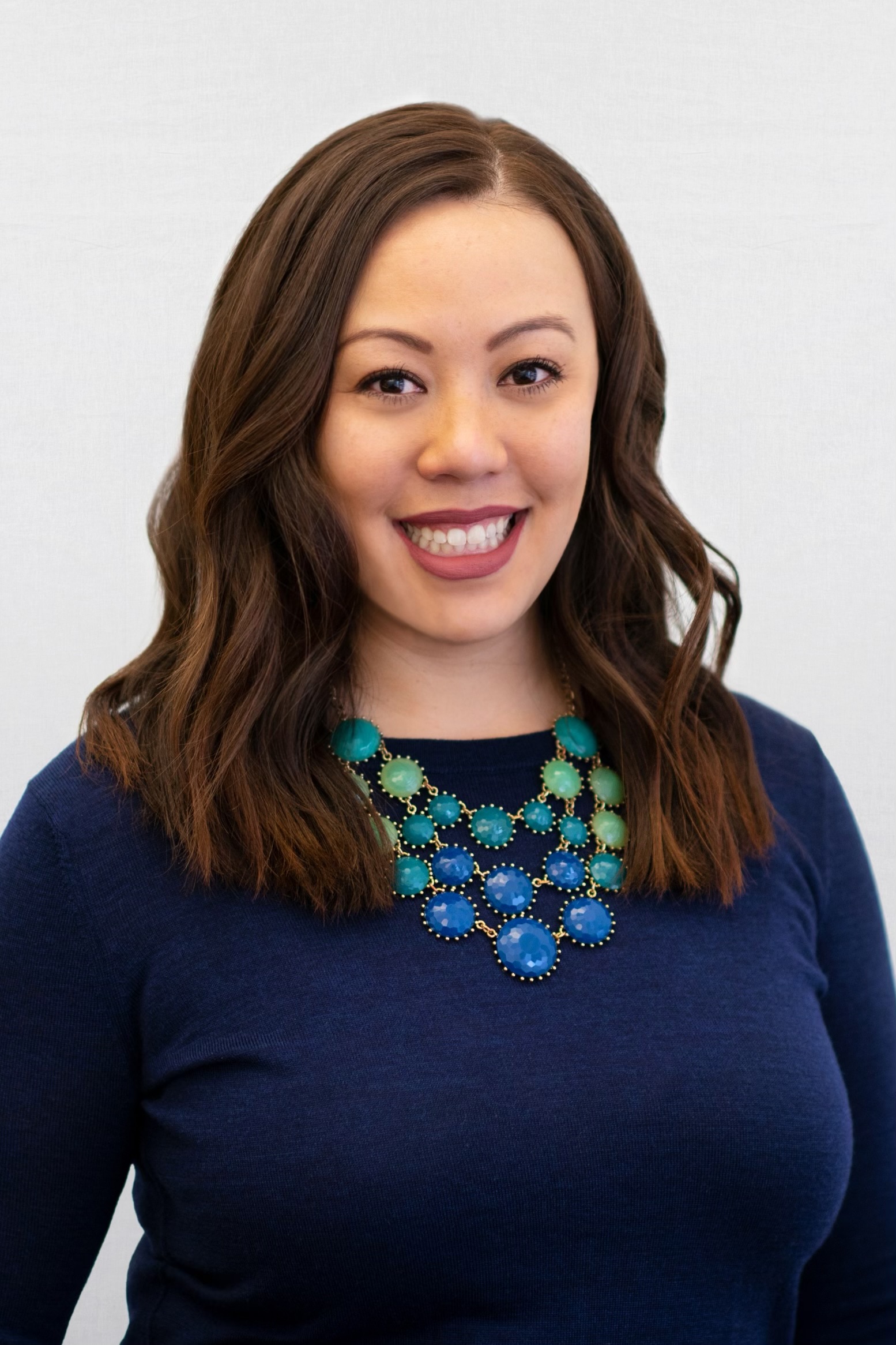 A smiling person of medium-light skin tone with long brown hair, wearing a blue sweater and blue-green chunky necklace.