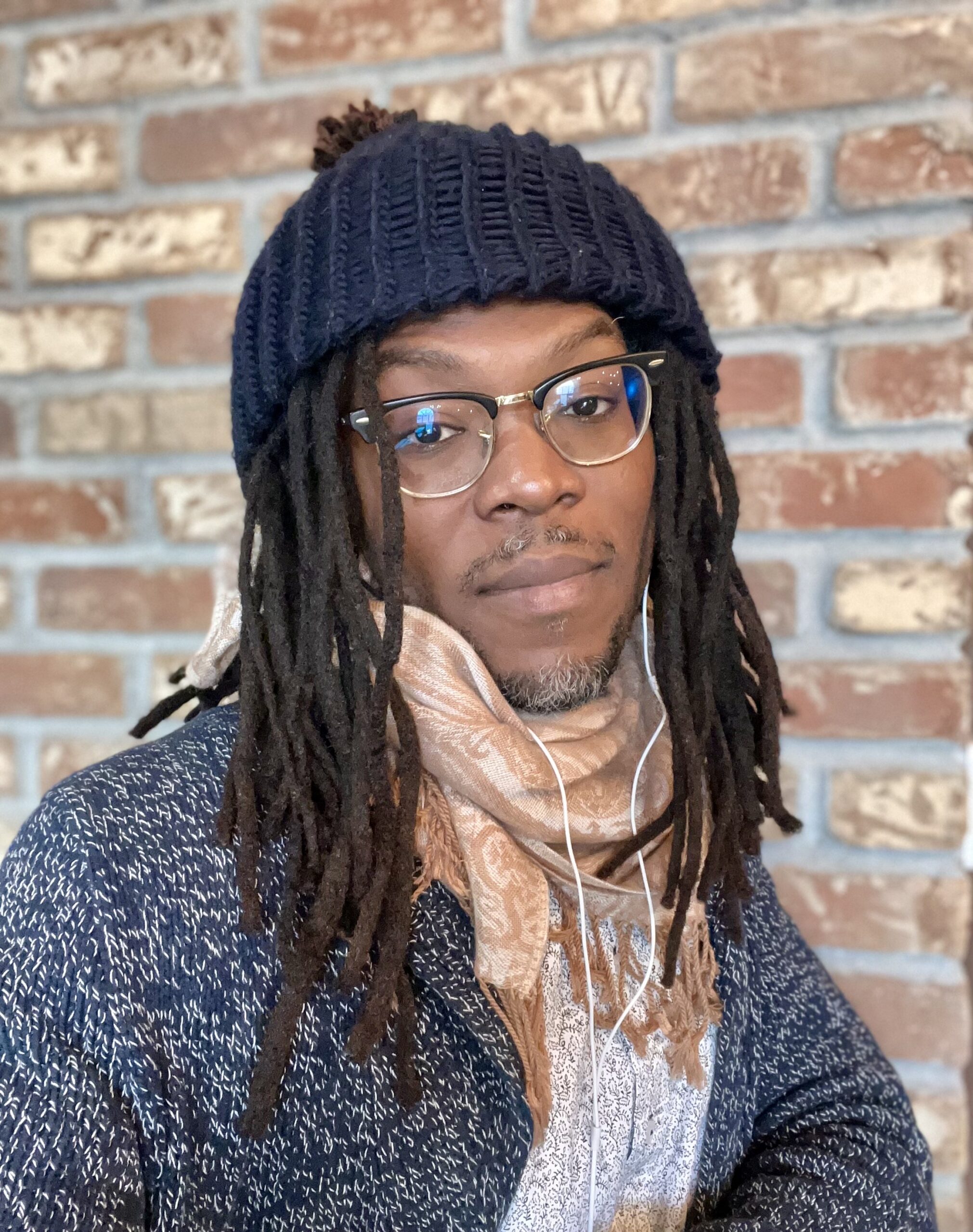 A person of medium-dark skin tone with long dreadlocs and glasses, wearing a navy blue beanie and sweater and orange scarf.