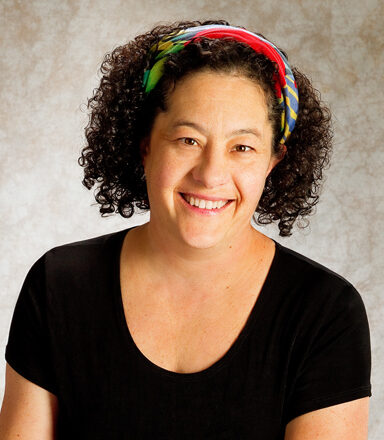 A person of light skin tone with dark, shoulder-length curly hair smiling. They are wearing a headband and a black tshirt.