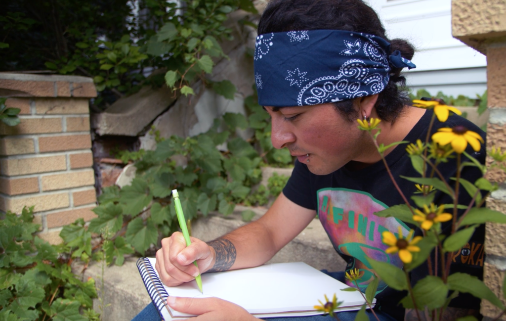 Medium-skin-toned person wearing a black tee and a blue bandana sits outside and sketches on a notepad.