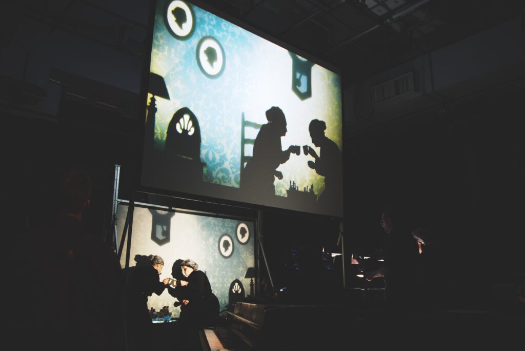 A projected image showing two silhouetted figures on a large screen on stage as two similar figures and set scene are under it.