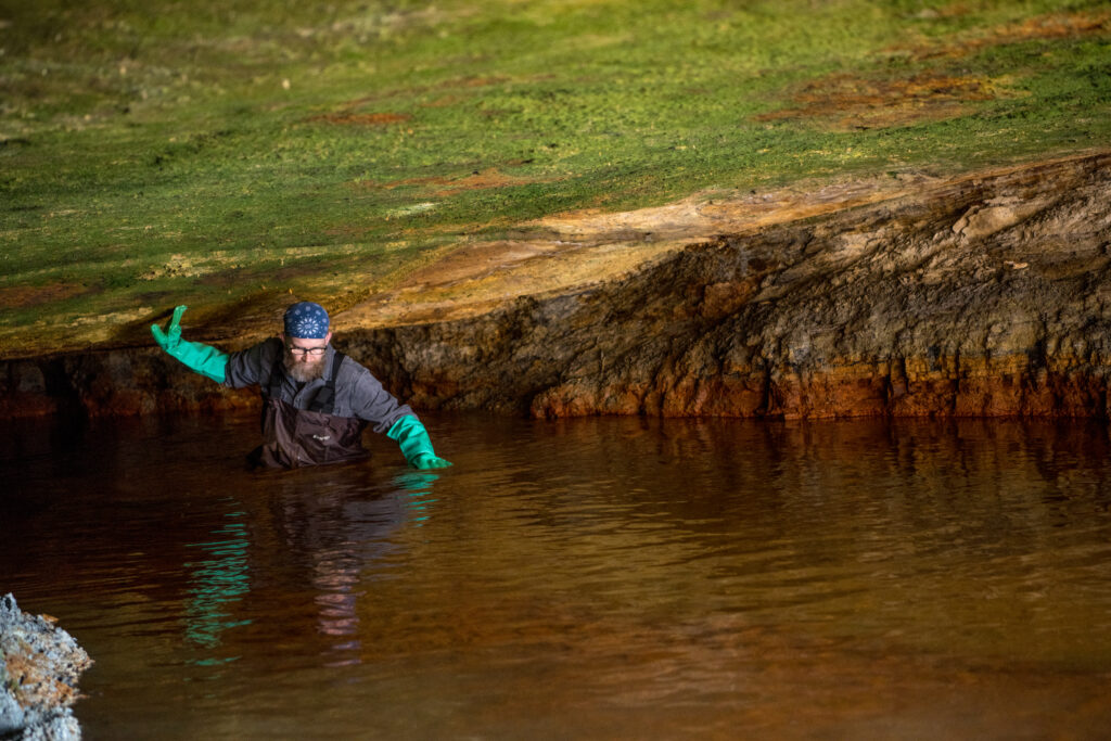 A light-skin-toned man with long gloves wades through a cavern of water and supports himself with his arm.