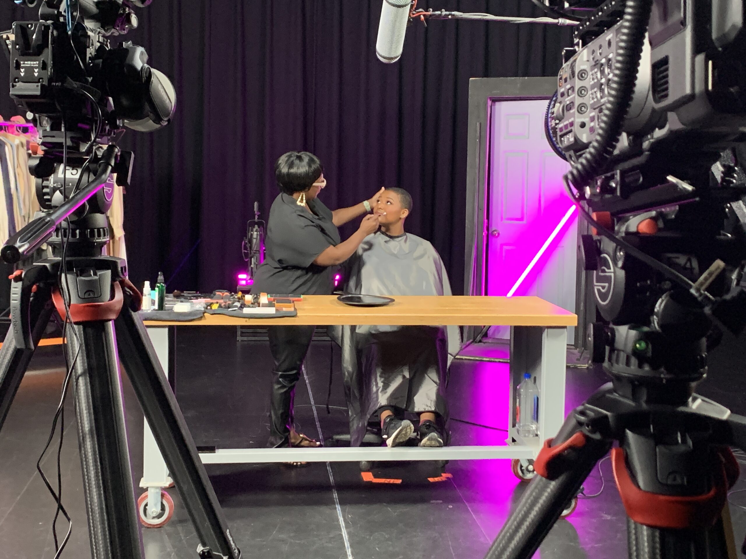 One dark-skinned woman applied makeup to another sitting down in front of large television cameras and a purple backdrop.