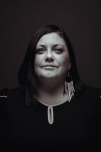 A grayscale headshot of a person with long dark hair, wearing a black blouse and beaded earrings.