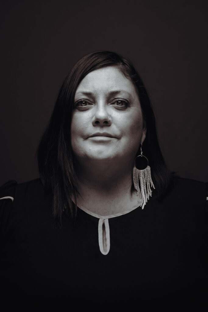 A grayscale headshot of a person with long dark hair, wearing a black blouse and beaded earrings.