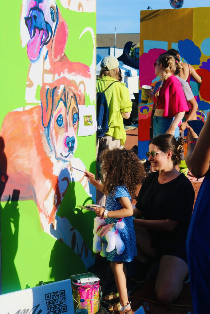 An adult helps a young child paint a mural of a dog at an outdoor event.