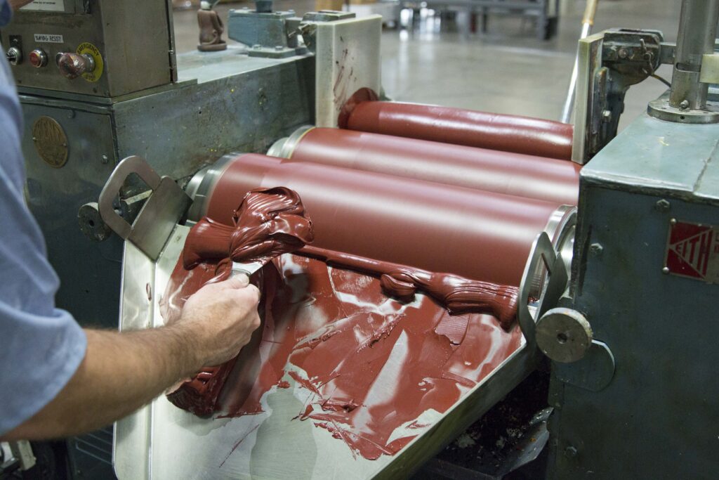 A hand works with dark red pigment material on a large roller machine.