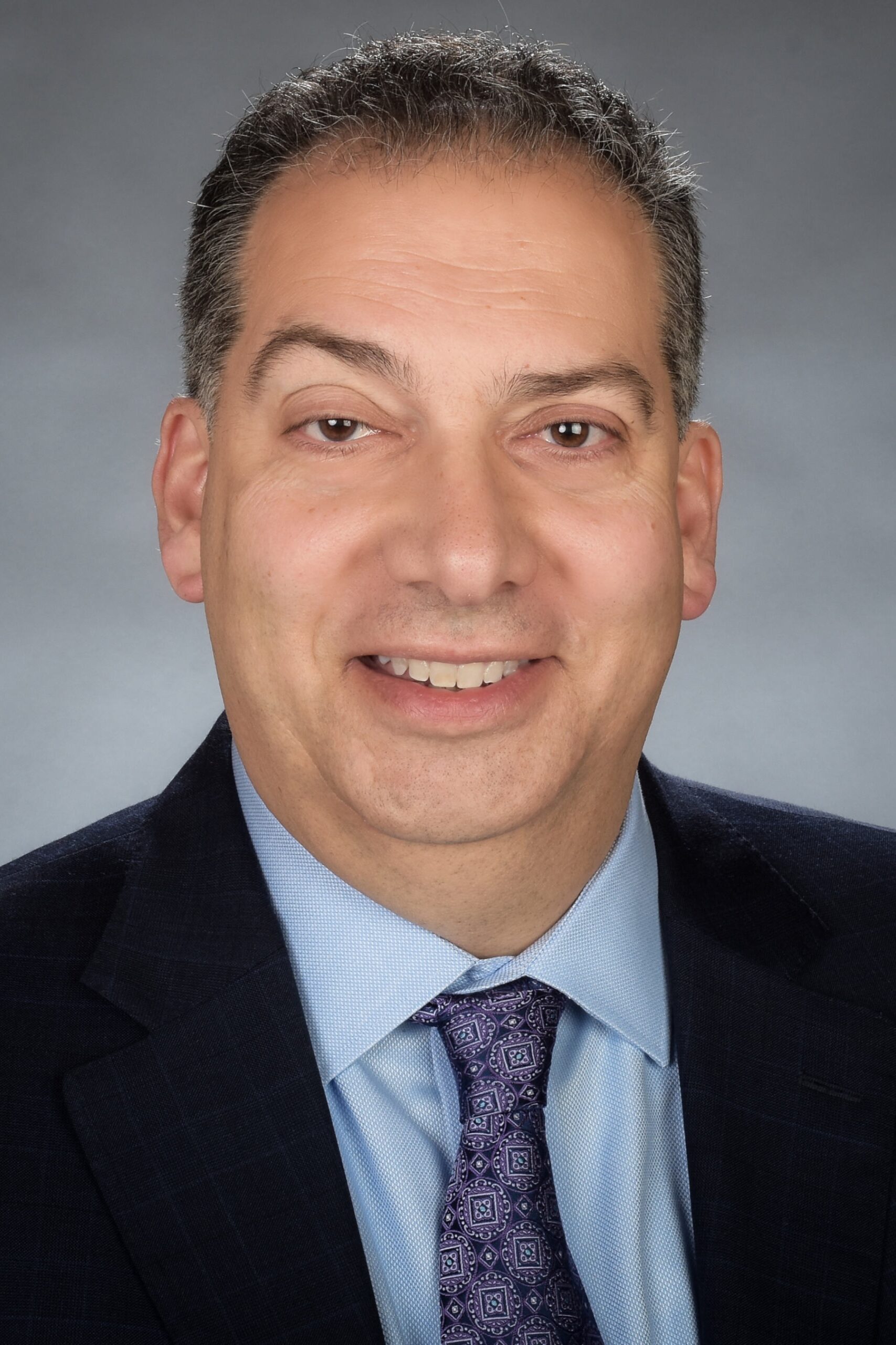 Headshot of a smiling person of medium light skin tone, short peppered hair, wearing a light blue shirt under a black suit jacket with a purple patterned tie.