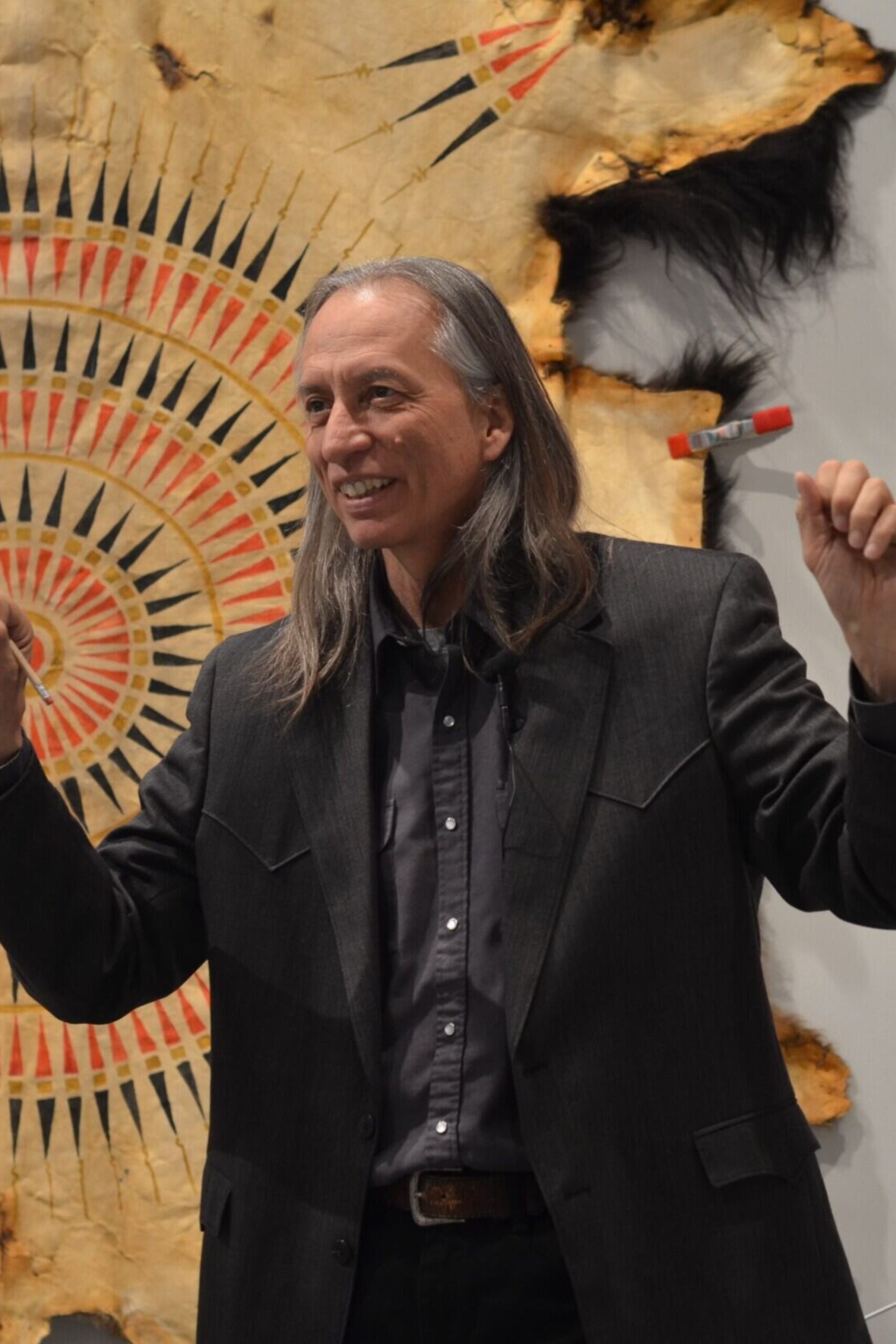 Close up of Craig Howe, speaking with a smile and both arms up, holding a pencil in one hand.