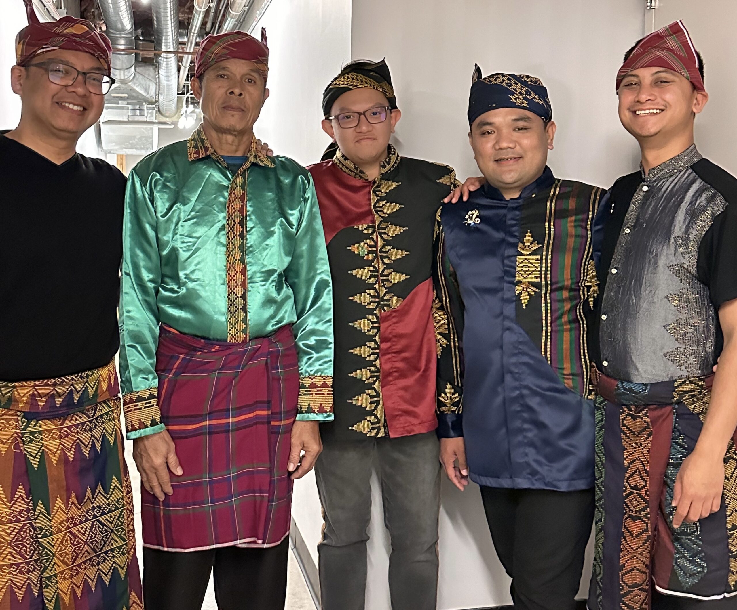 Five smiling people of medium skin tone wearing traditional hand-woven "Inaul" clothing.