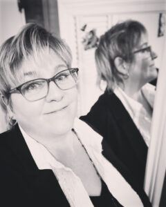 Black and white photo of a person with short blonde hair smiling next to a mirror that partially reflects their image.