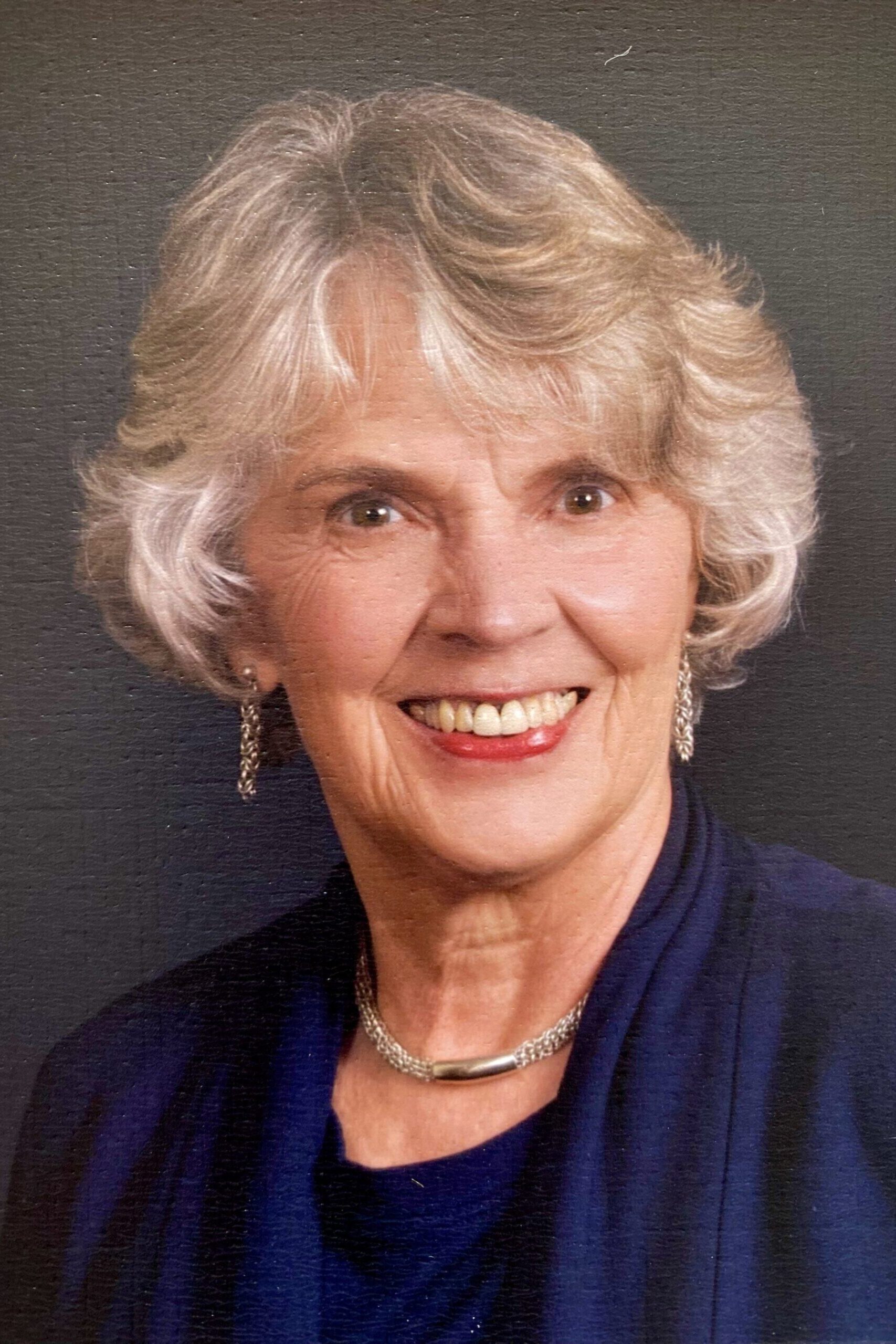 Headshot of a smiling person of light skin tone with short brown hair, wearing dangling earrings and cream and black wool blazer.