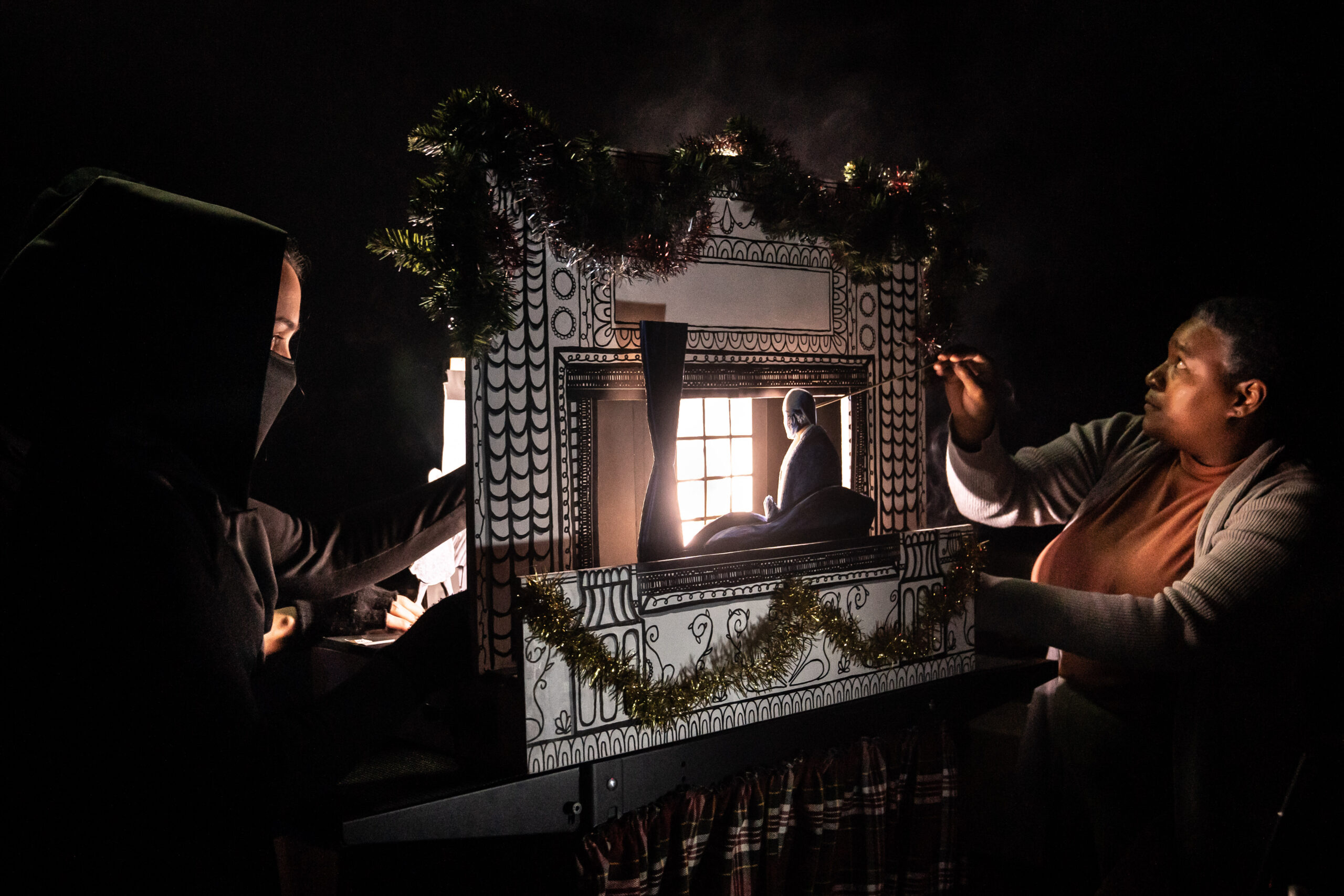 Two people standing by a miniature stage set as they operate paper puppets with their hands.