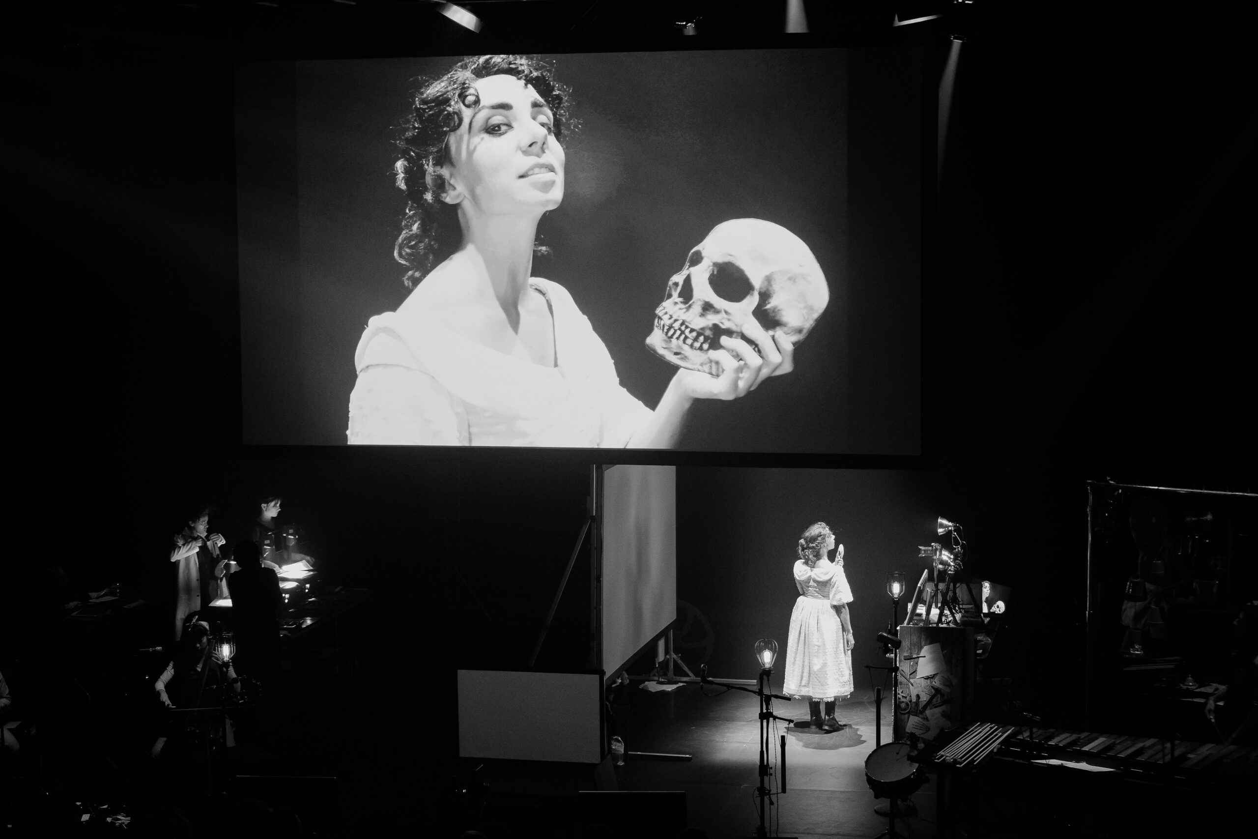 A projected image of a person holding a skull-shaped mask in one hand. The person is standing under the large projected image on stage.