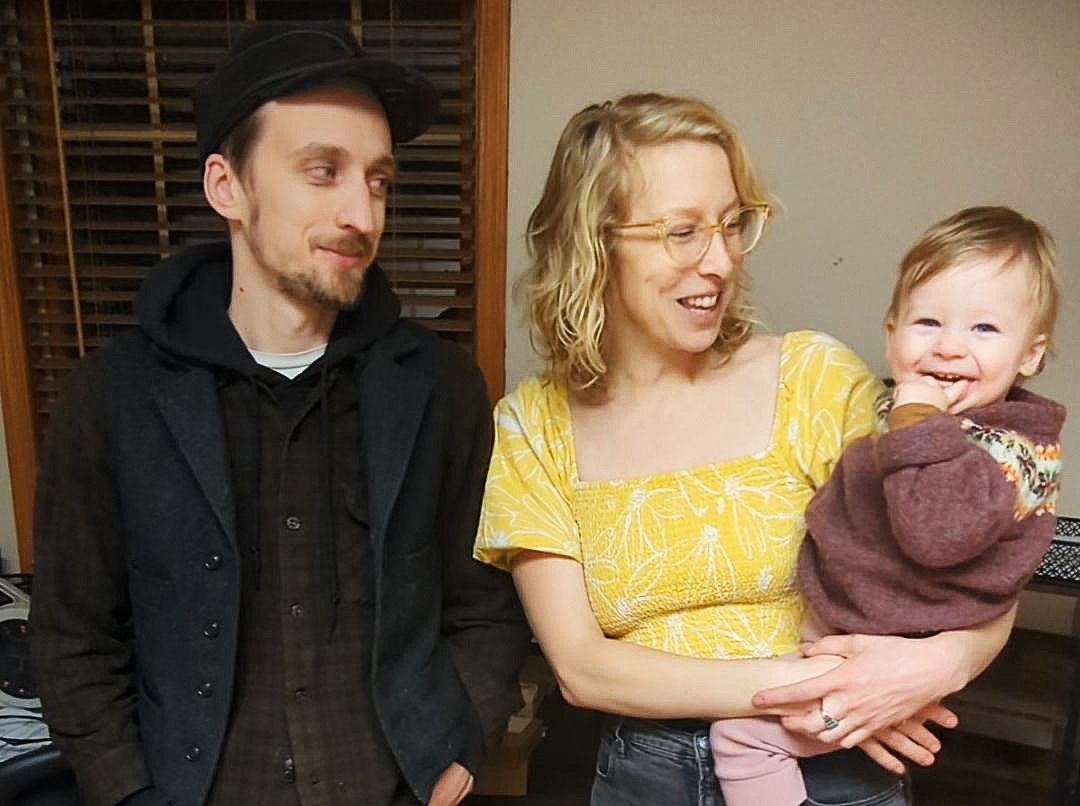 Light-skinned man, woman and baby smile inside a room.