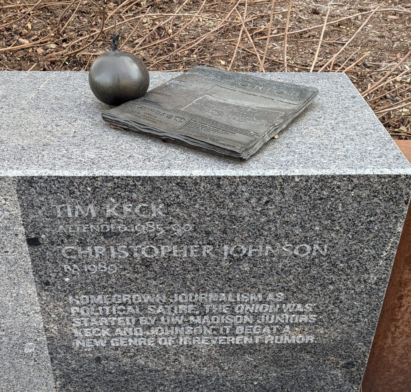 A stone block sculpture with engraved text with carved sculptures depicting an onion and a newspaper resting on top of it.