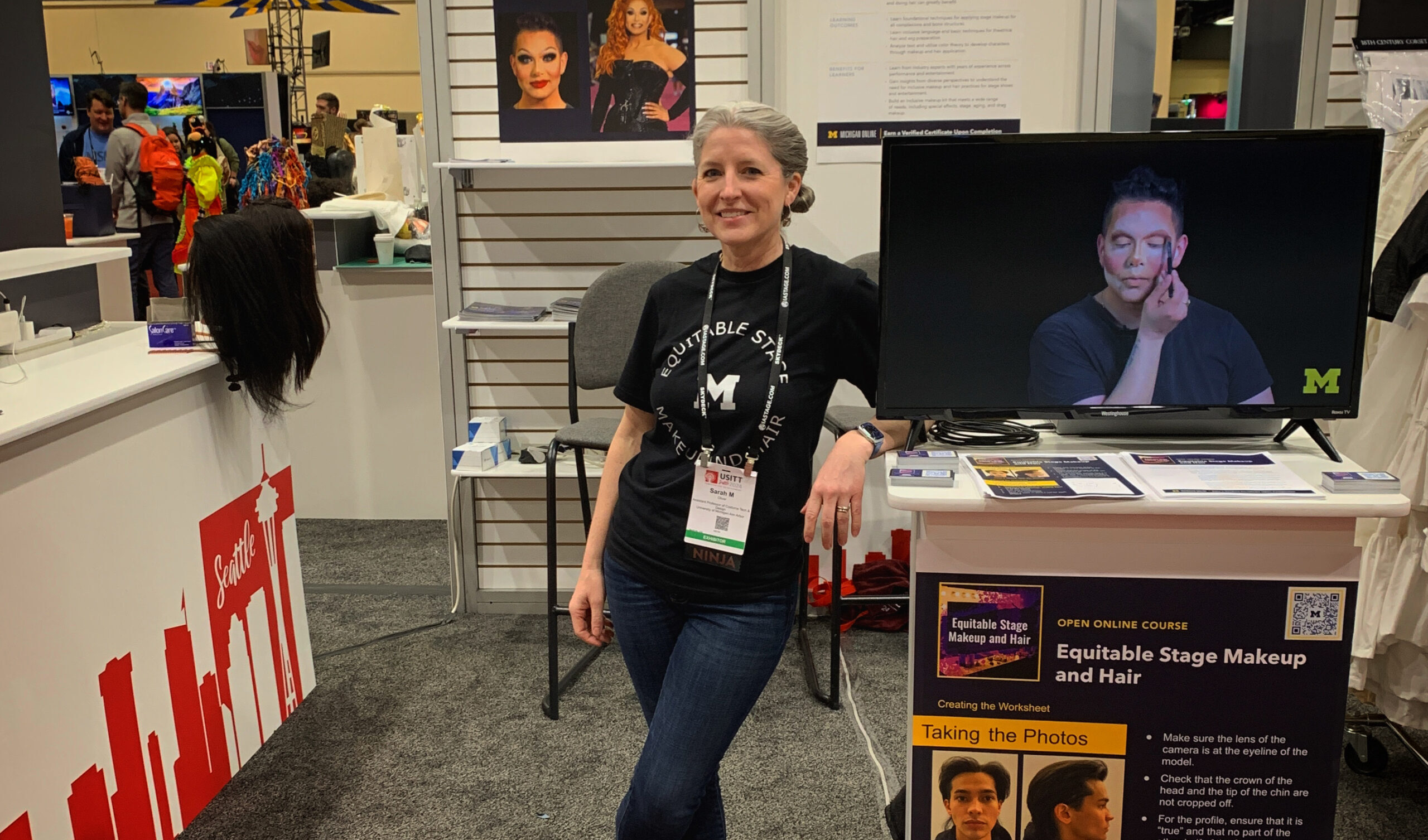 Light-skinned woman stands next to television screen of a person applying makeup.