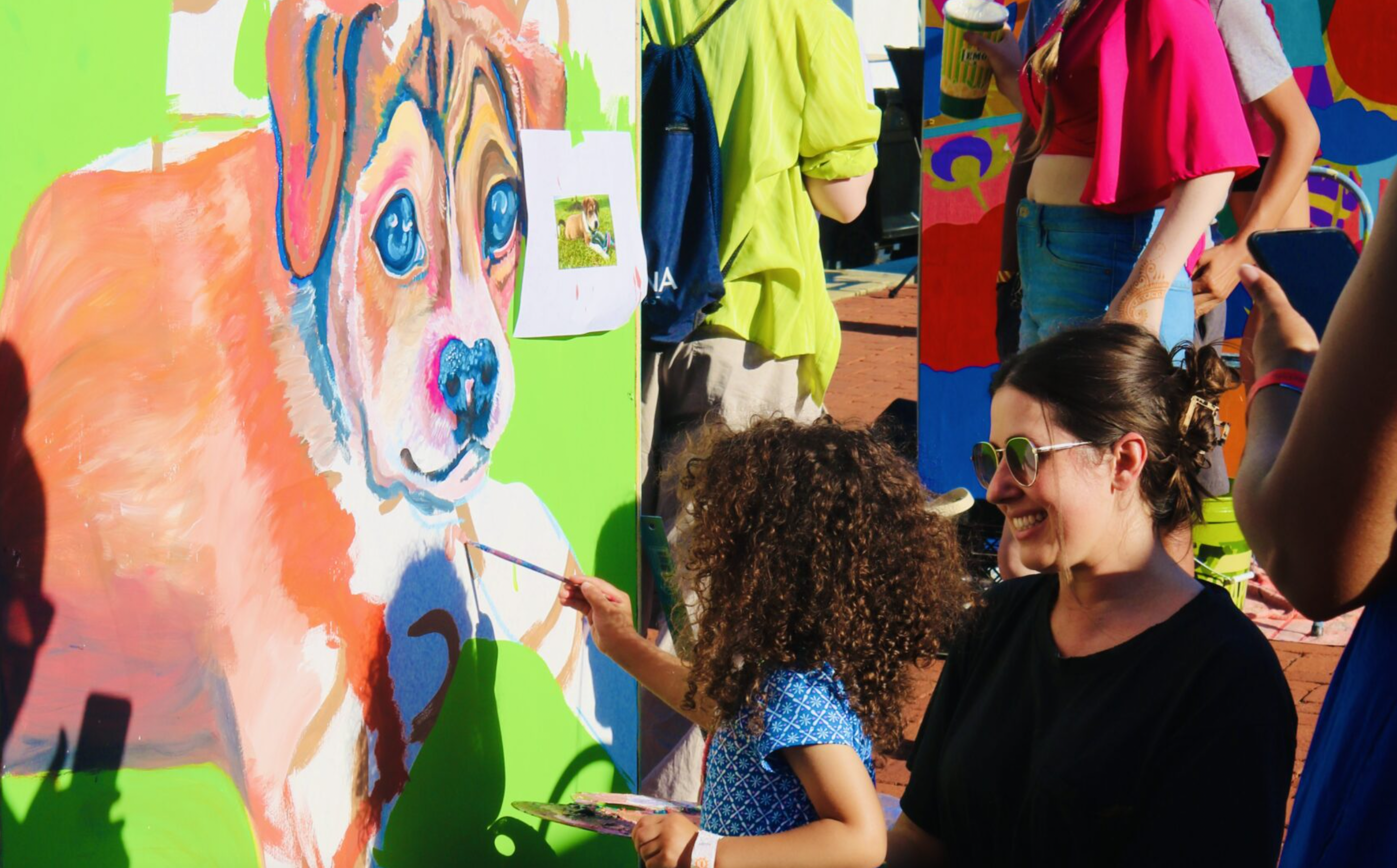 An adult helps a young child paint a mural of a dog at an outdoor event.