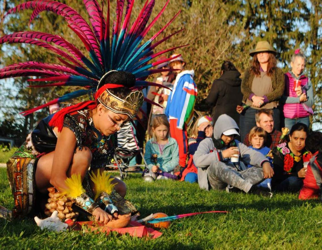 A person wearing a headgear with colorful feathers and traditional clothing for dance is kneeling and holding a bowl-like object placed on the ground. They are outdoors with other people around watching them.