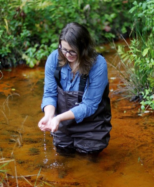 Light-skinned woman cups hands with water while standing in an orange creek.