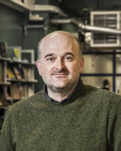 Headshot of a bald man with light skin wearing a green sweater, with bookcases behind him.