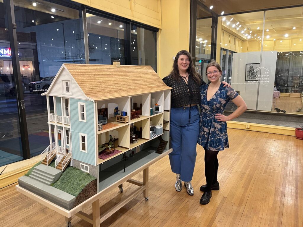 Two smiling people pose next to a miniature house in an art gallery