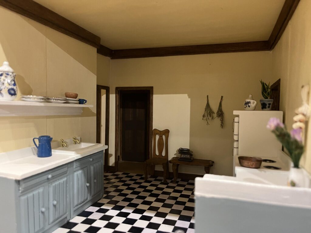 A miniature recreation of a kitchen, with a sink, fridge, and stove, with blue cabinets and checkerboard flooring.